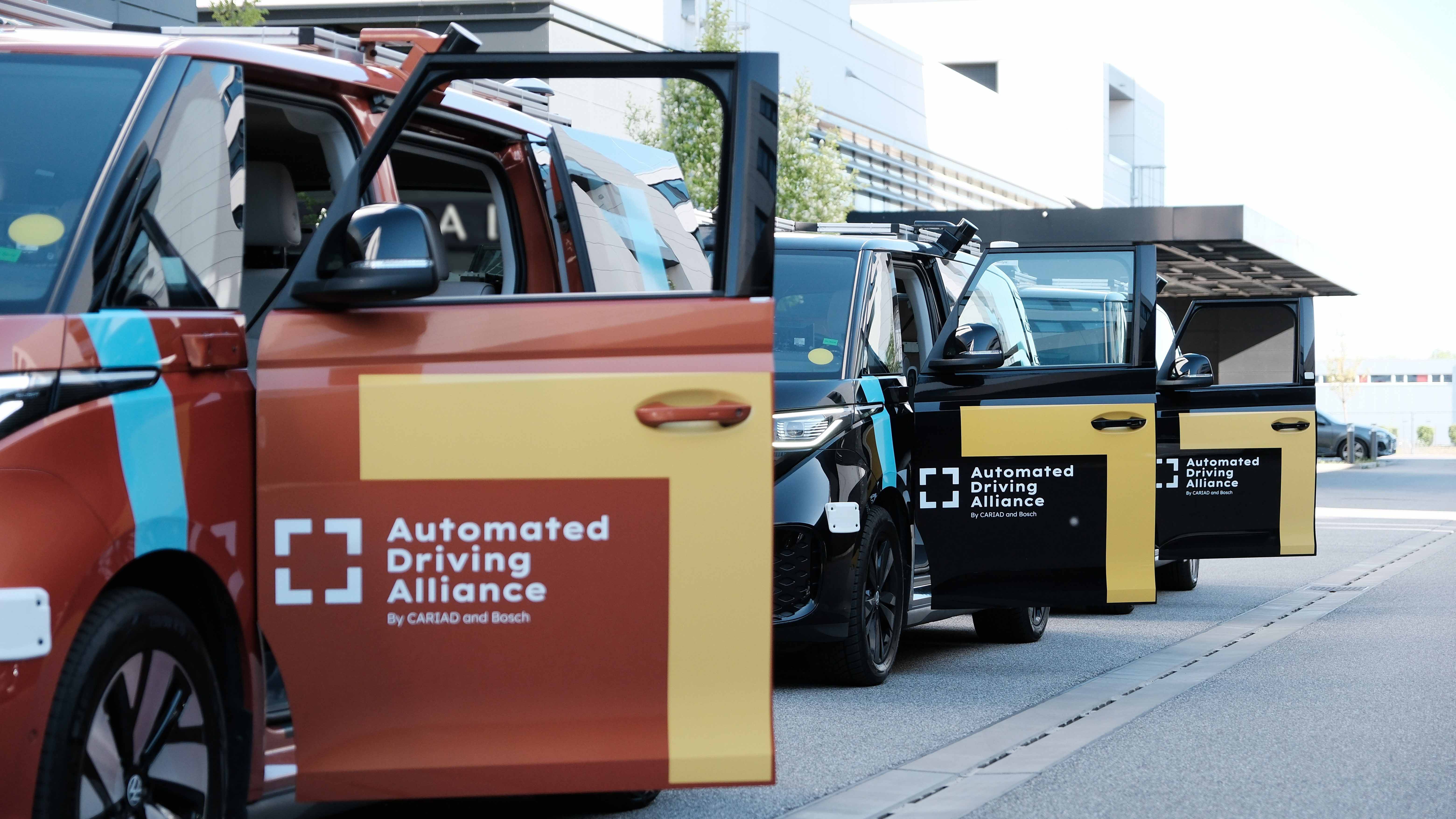 Multiple sensor-equipped test vehicles from the Automated Driving Alliance with open doors parked outside a modern office building, showcasing autonomous driving technology.