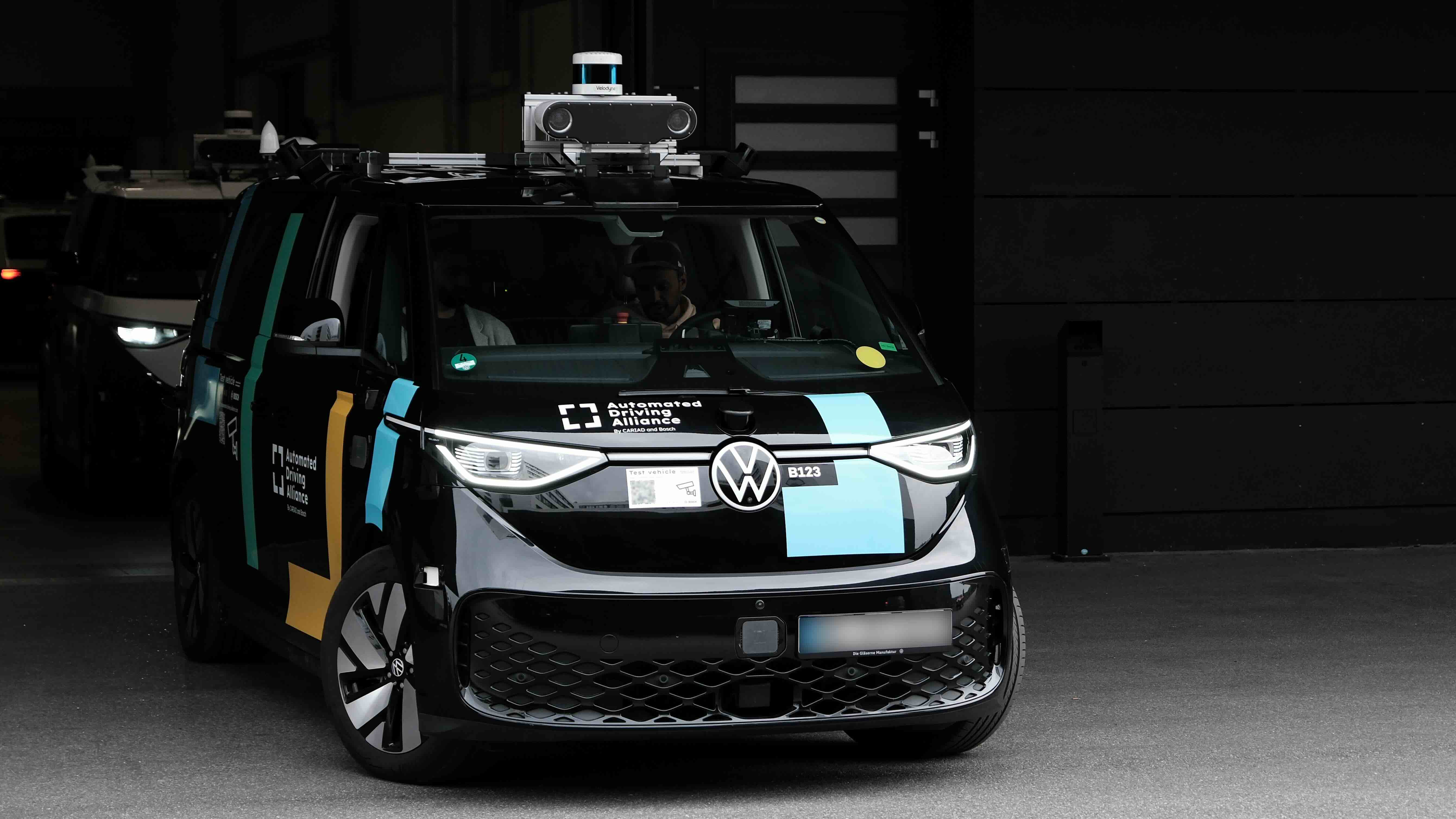Black Volkswagen ID.Buzz with roof-mounted sensors and cameras, branded with 'Automated Driving Alliance', driving out of a workshop