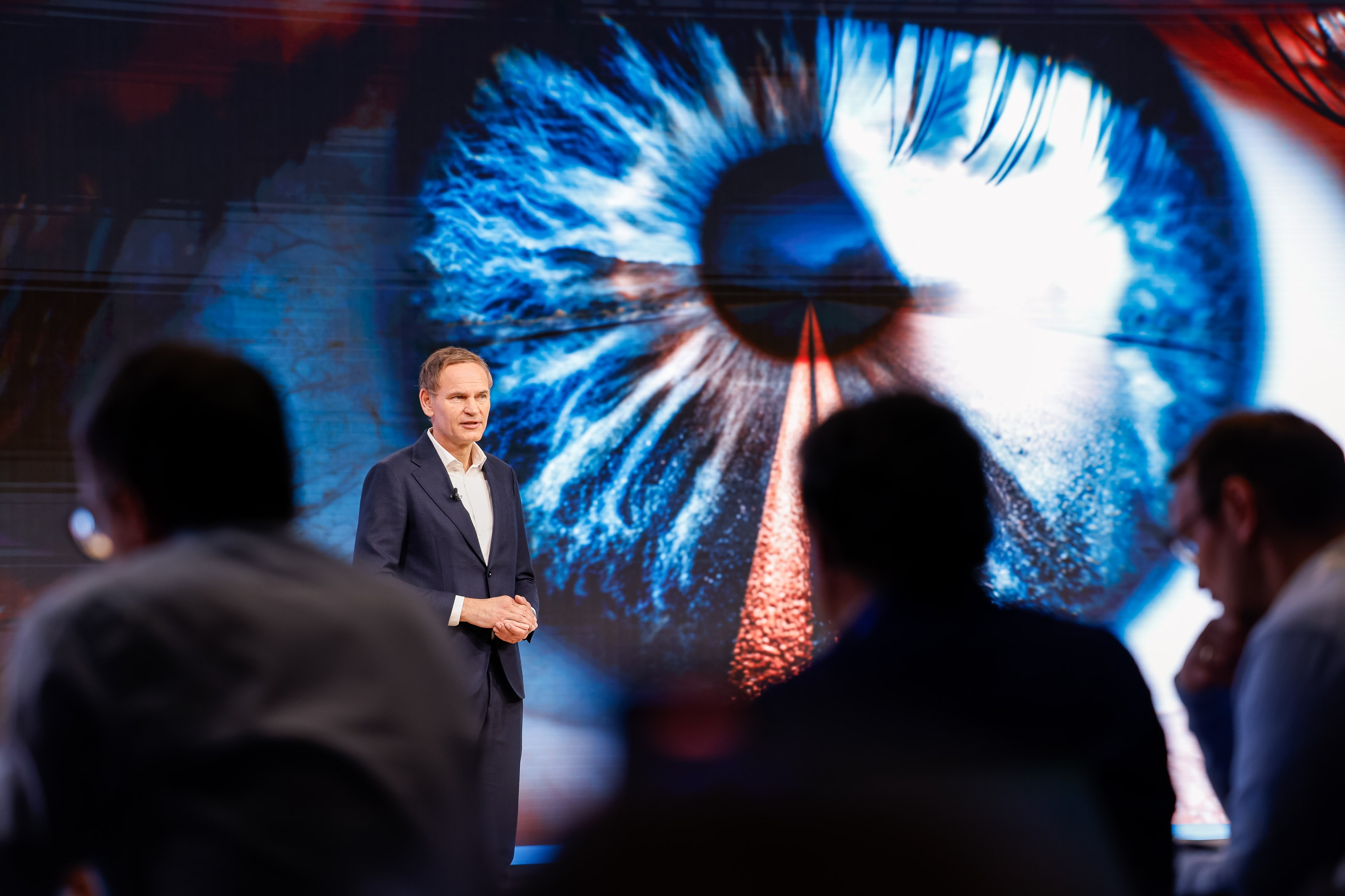 Oliver Blume standing on a stage in front of a large, high‑resolution visual of an eye displayed on an LED screen, addressing an audience in a conference or keynote setting.