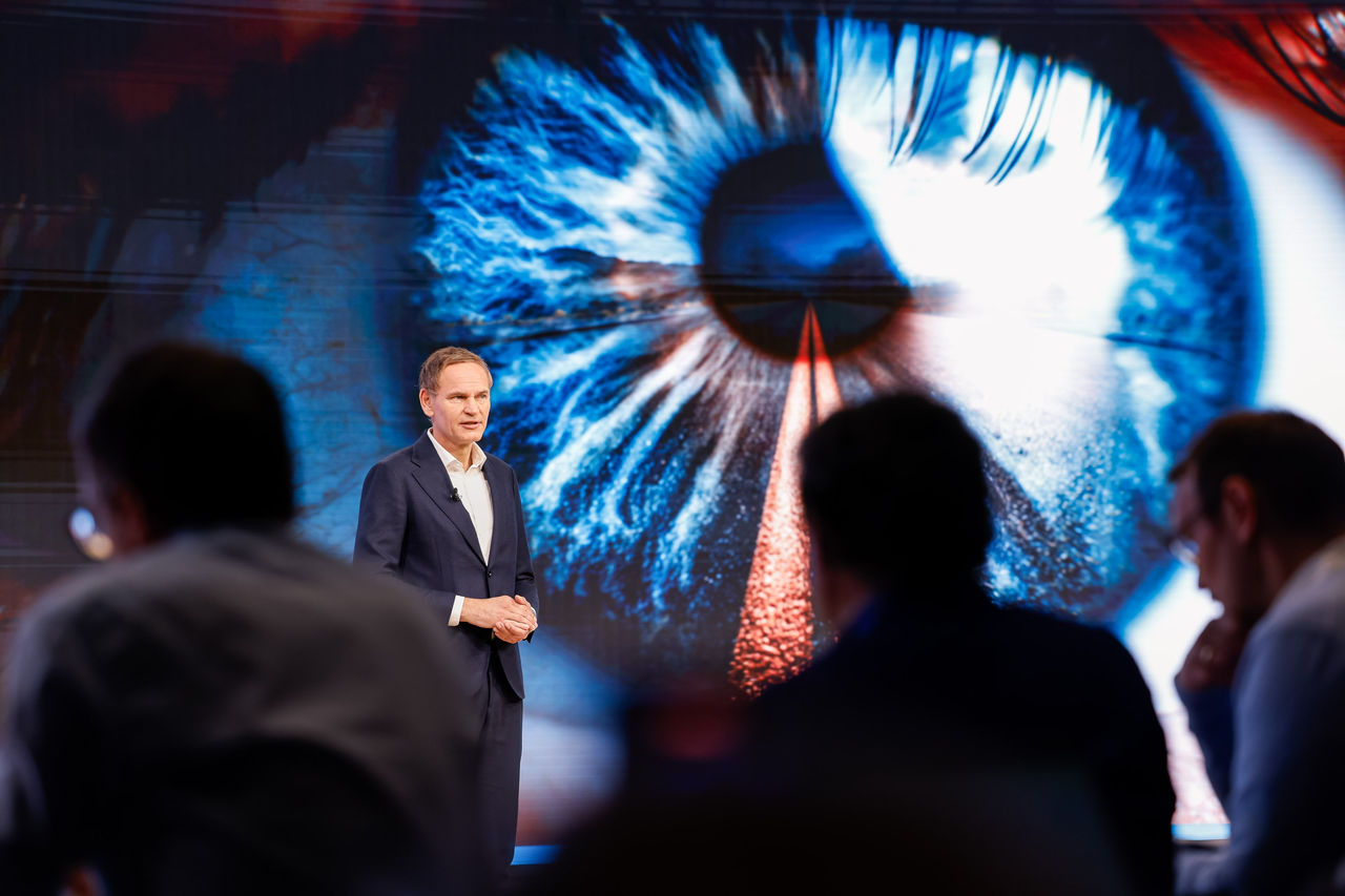 Oliver Blume standing on a stage in front of a large, high‑resolution visual of an eye displayed on an LED screen, addressing an audience in a conference or keynote setting.