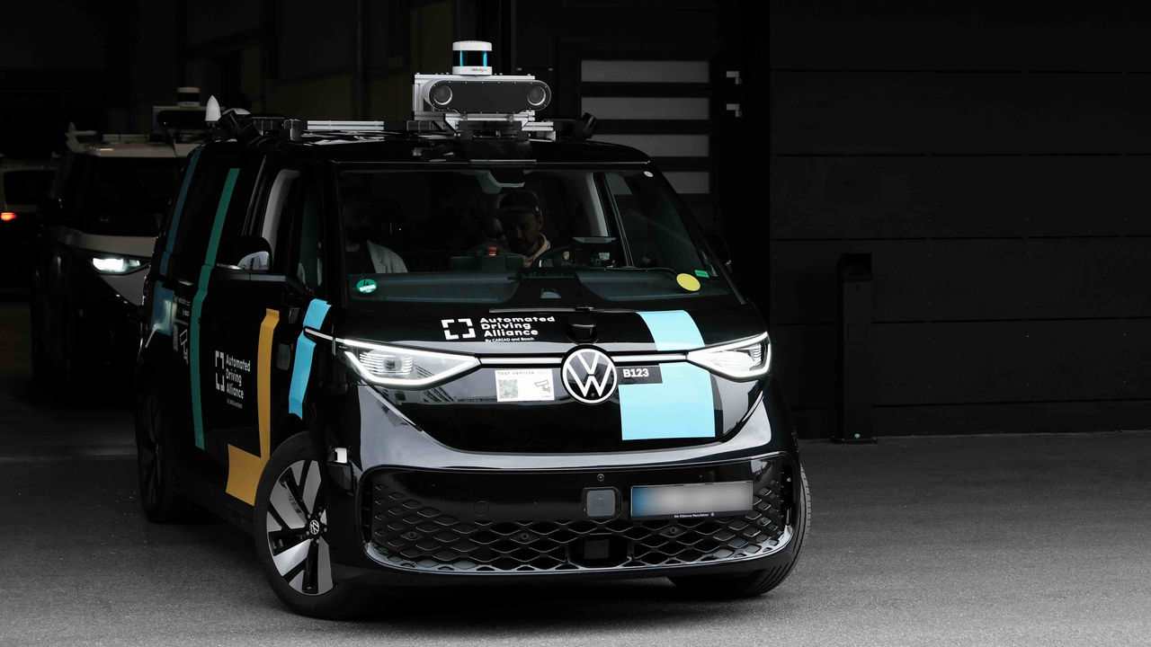 Black Volkswagen ID.Buzz with roof-mounted sensors and cameras, branded with 'Automated Driving Alliance', driving out of a workshop