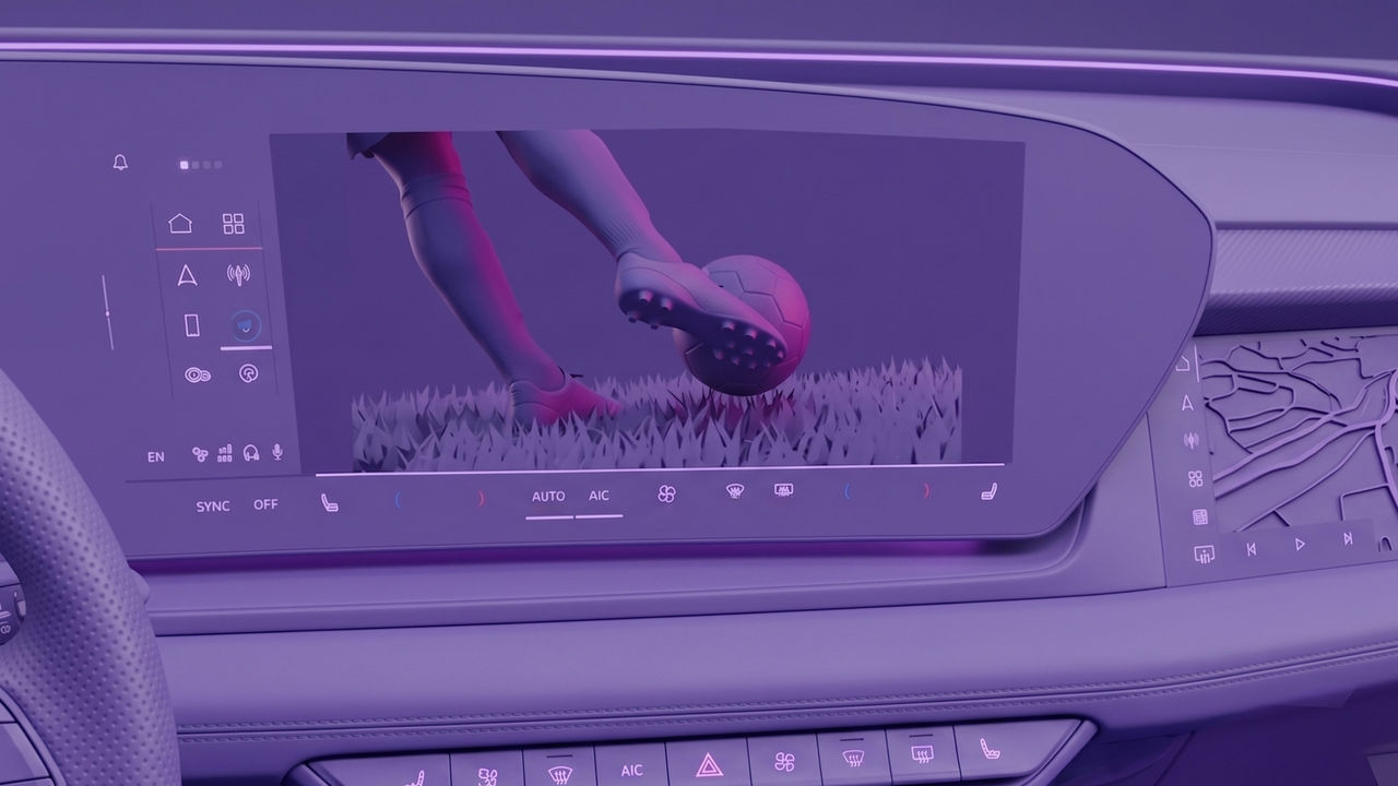 A modern car interior featuring a wide digital infotainment display showing a live football scene with a player kicking a ball on a stadium pitch. The dashboard integrates a sleek, high-resolution touchscreen with illuminated user interface icons for navigation, apps, connectivity, and vehicle functions. The cockpit design highlights ambient lighting in purple and blue tones, a premium textured dashboard surface, and climate control indicators below the screen. The steering wheel is partially visible in the foreground, reinforcing the in-car perspective. The image illustrates connected vehicle software enabling entertainment and real-time content during a journey, blending automotive UX design with sports streaming in a smart mobility context.